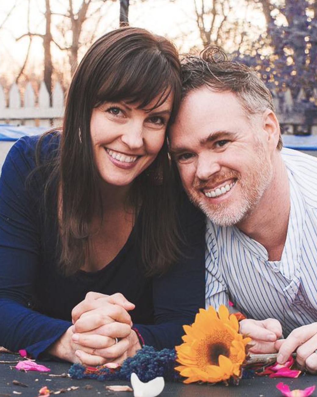 Ashley and Drew Robinson rest side by side outdoors at golden hour, smiling toward the camera with their hands clasped beside a sunflower.