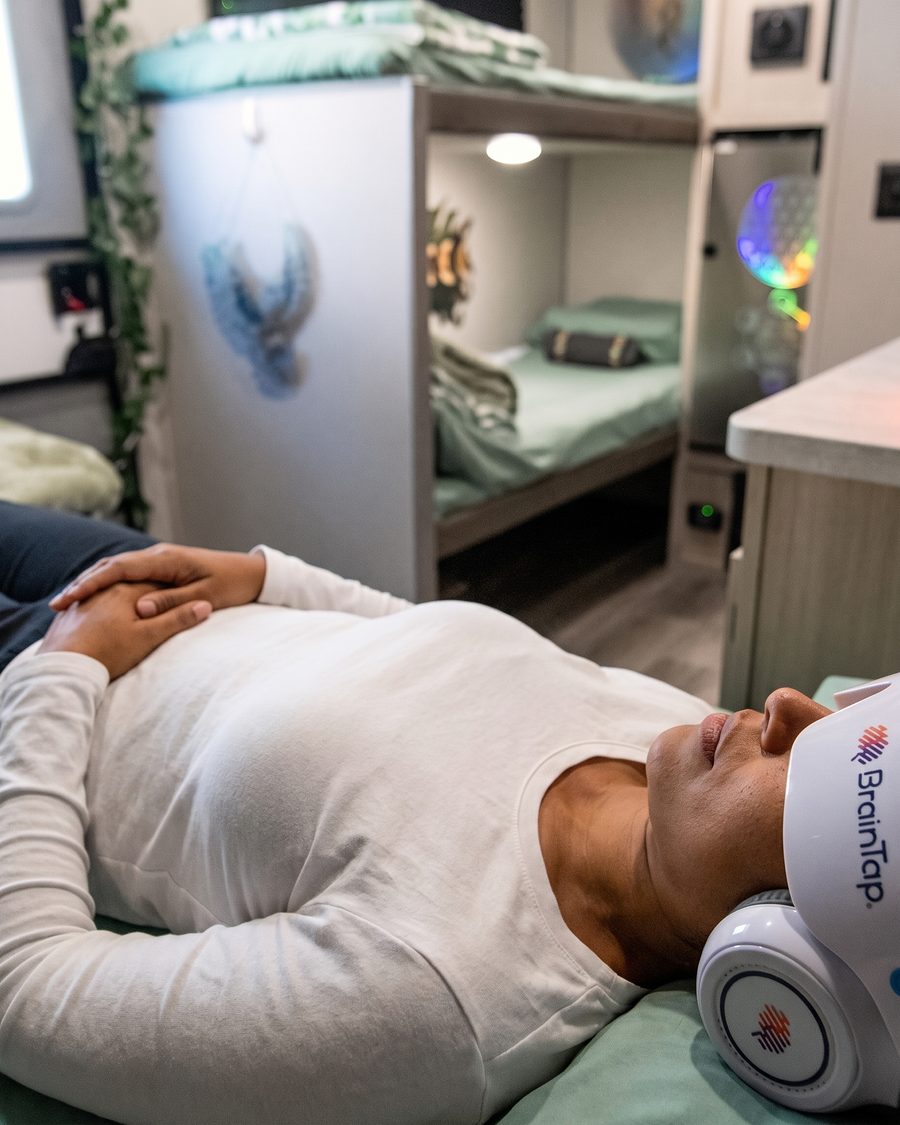 A guest in a white shirt rests mid-session with hands folded, the BrainTap headset over her eyes, the Pod’s bunks soft behind her.